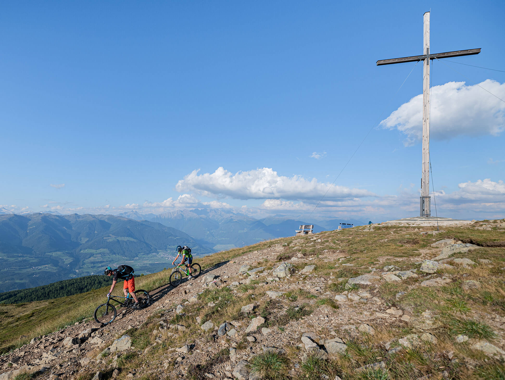 Zwei Mountainbiker treten die Abfahrt an - daneben ein Gipfelkreuz - Hotel Montis