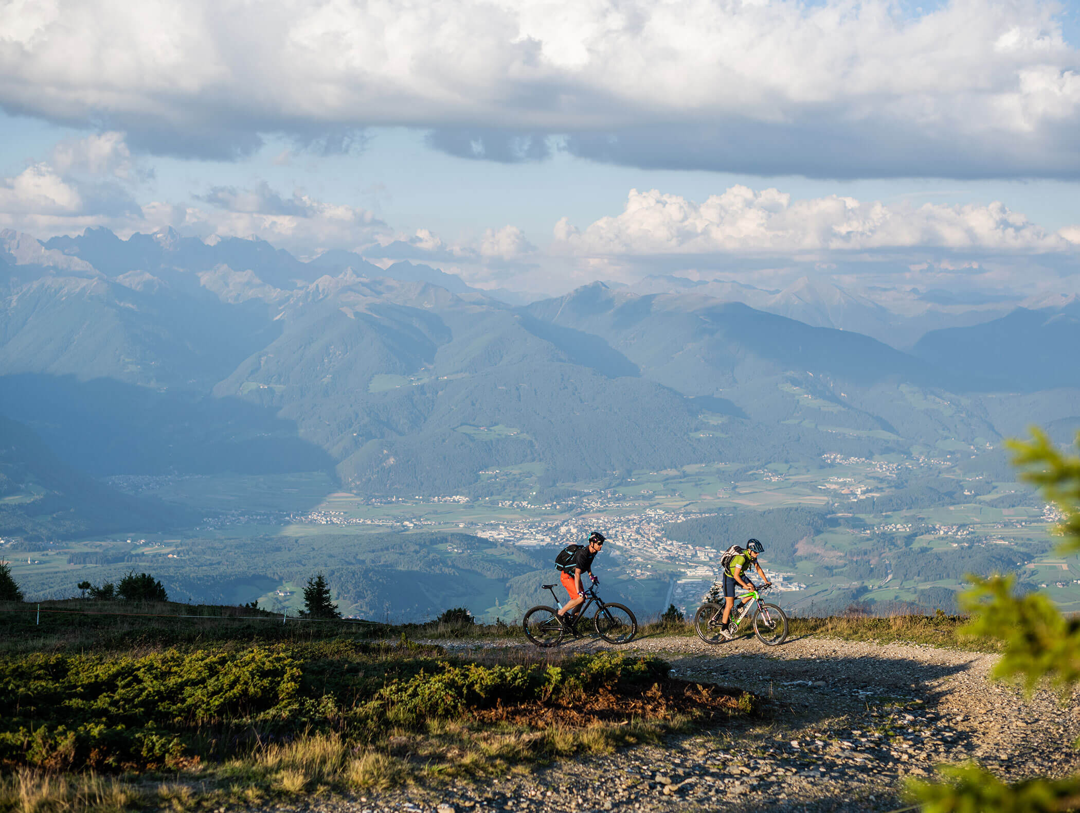 Zwei Mountainbiker auf einem Forstweg, dahinter das Tal mit der Stadt Bruneck - Hotel Montis