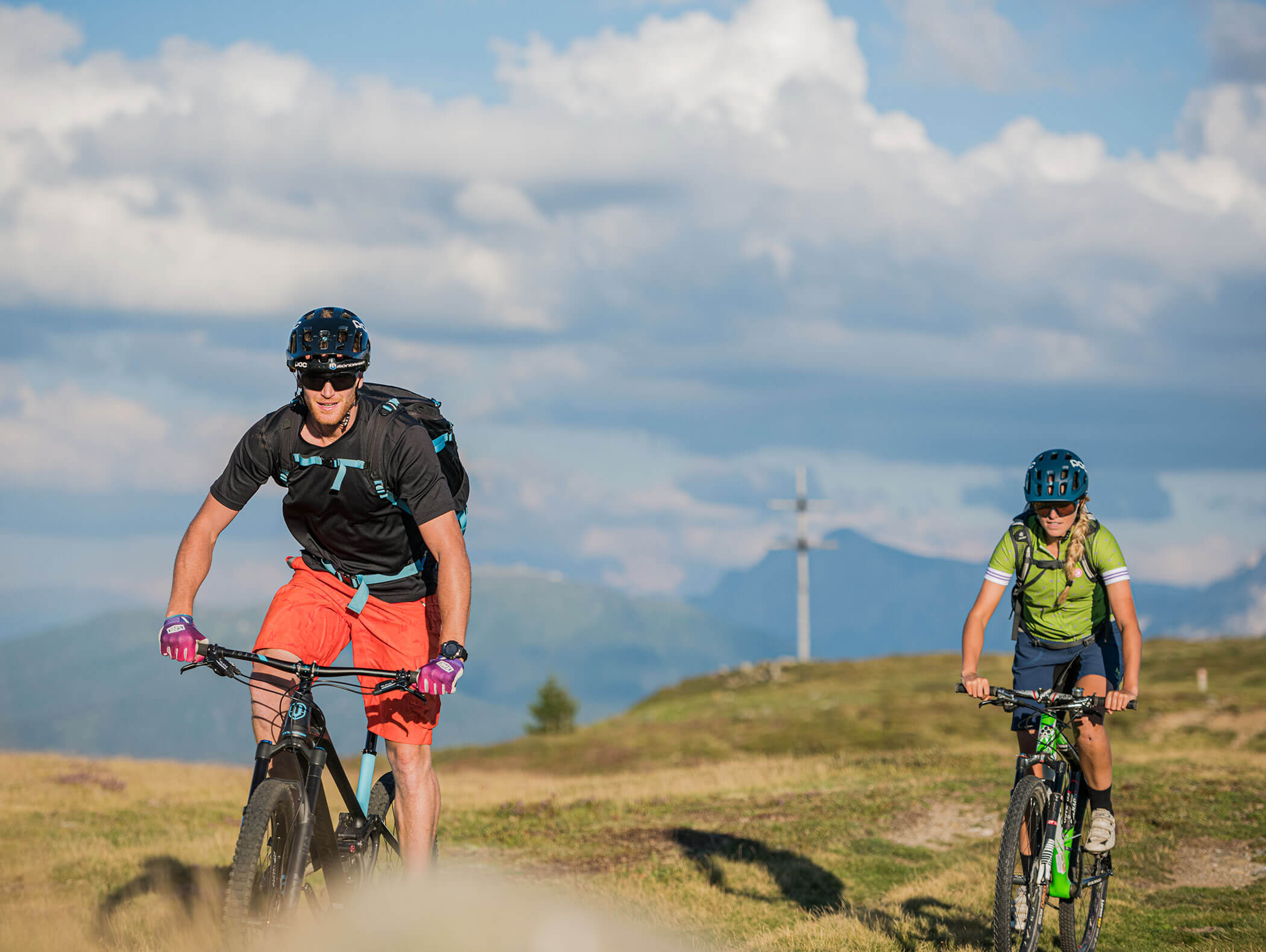 Ein Pärchen beim Mountainbiken, dahinter ein Gipfelkreuz - Hotel Montis