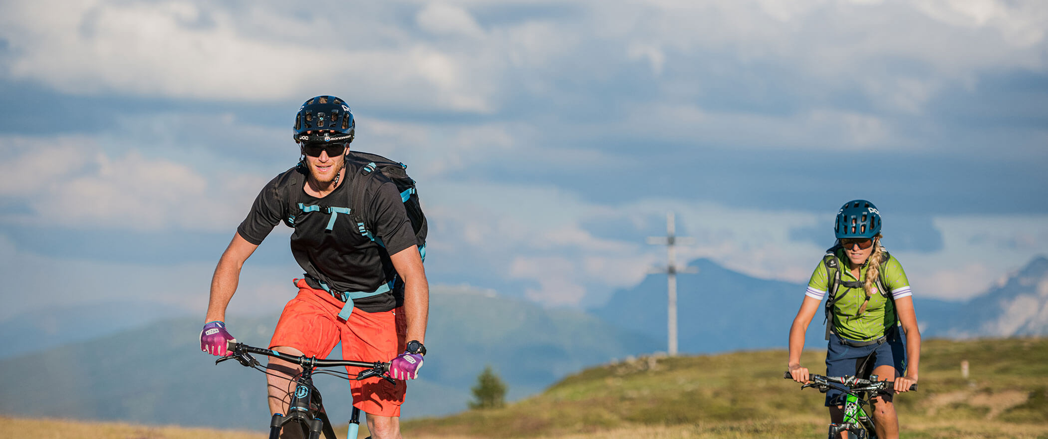 Ein Pärchen beim Mountainbiken, dahinter ein Gipfelkreuz - Hotel Montis