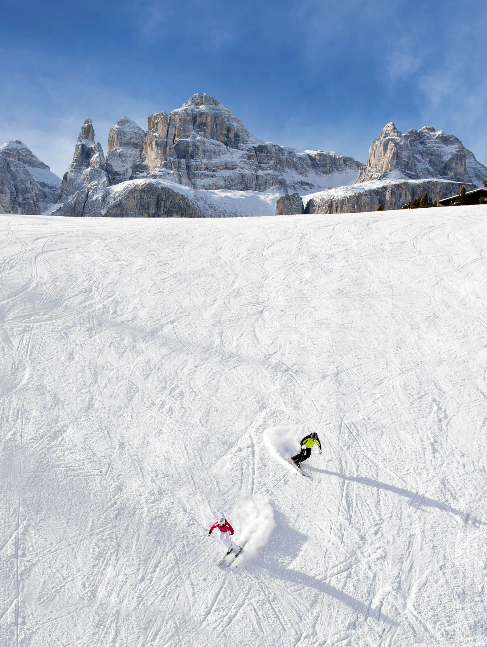 Zwei Skifahrer fahren die Piste hinunter, dahinter die Bergspitzen der Dolomiten - Hotel Montis